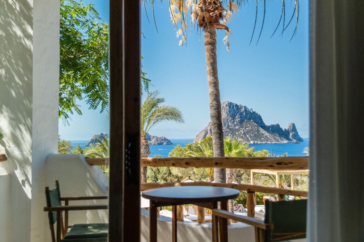 Vue pittoresque du balcon avec chaises, table, palmiers et le rocher emblématique d'Es Vedrà sur la mer.