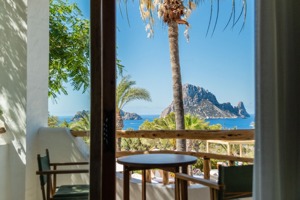 Vue pittoresque du balcon avec chaises, table, palmiers et le rocher emblématique d'Es Vedrà sur la mer.