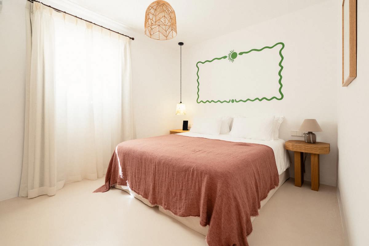 Chambre lumineuse avec couvre-lit rose, rideaux blancs, meubles en bois et cadre vert au-dessus du lit.