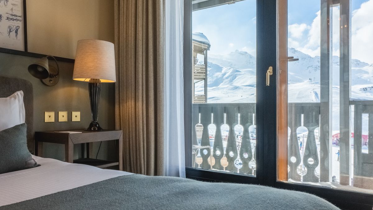 Table d'appoint avec lampe près du lit, vue sur montagnes enneigées à travers verre.