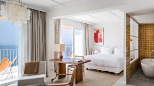 A bedroom features a cozy bed, a cozy chair by the table, and a glass window with a wonderful ocean view.