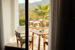 View through curtains onto a sunny terrace with wooden chairs, table, palm trees, and distant hills.
