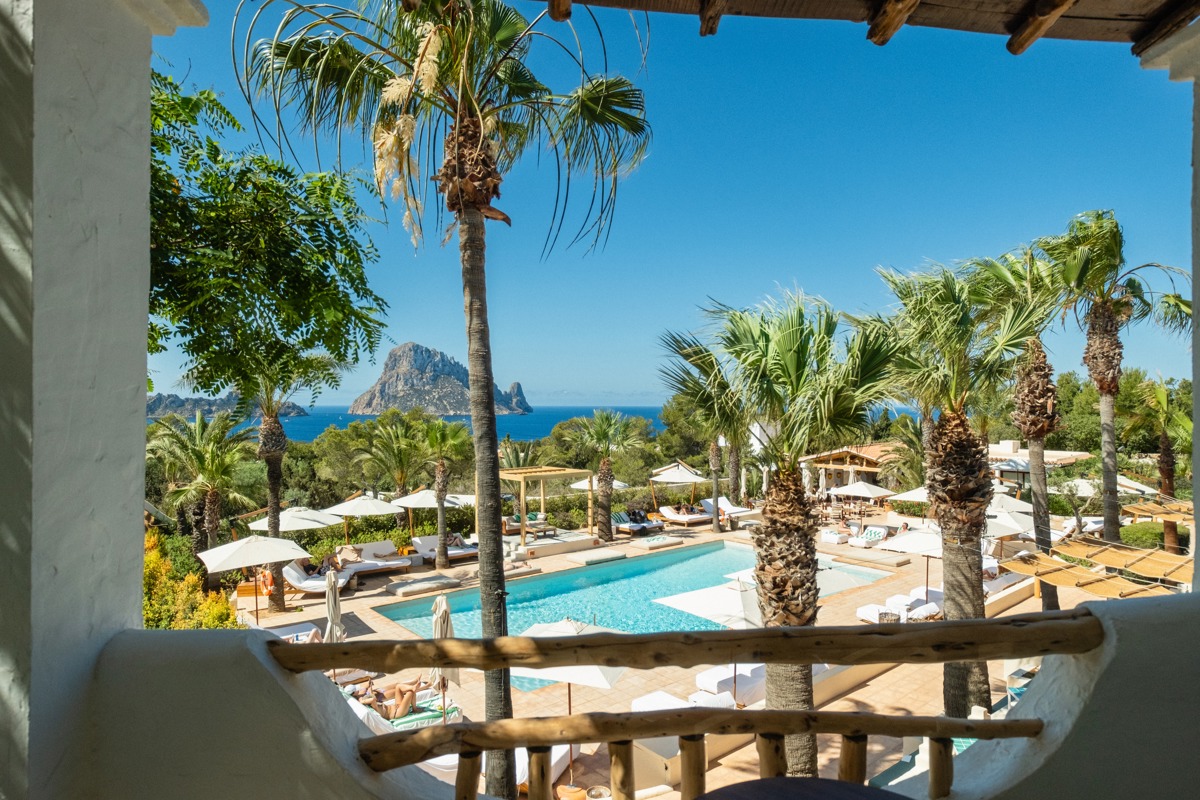 Vue du balcon sur palmiers, piscine de luxe, transats et île d’Es Vedrà sous ciel clair.