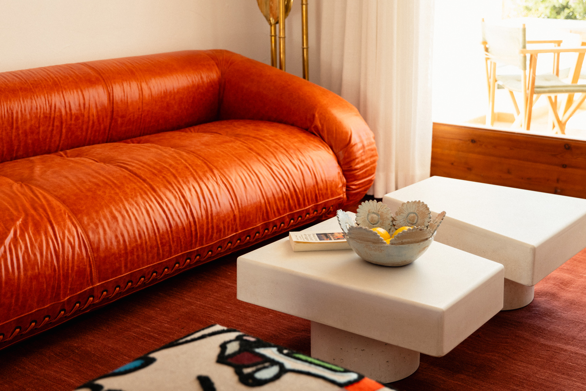 Salon confortable avec canapé en cuir orange, tables basses blanches, bol décoratif et terrasse ensoleillée.