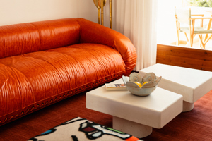 Salon confortable avec canapé en cuir orange, tables basses blanches, bol décoratif et terrasse ensoleillée.