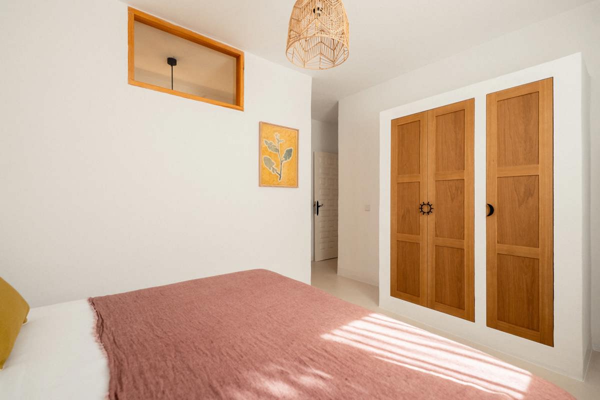 Chambre minimaliste avec couvre-lit rose, armoire en bois, suspension en osier et œuvre botanique encadrée.