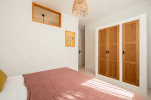 Chambre minimaliste avec couvre-lit rose, armoire en bois, suspension en osier et œuvre botanique encadrée.