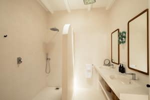 Salle de bain minimaliste avec douche italienne, double vasque, grands miroirs et murs beige clair éclairés naturellement.