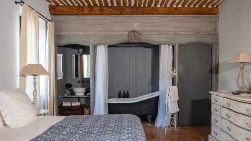 A tidy bedroom features a cozy bed, side table with lamp, shelves, and visible bathtub.