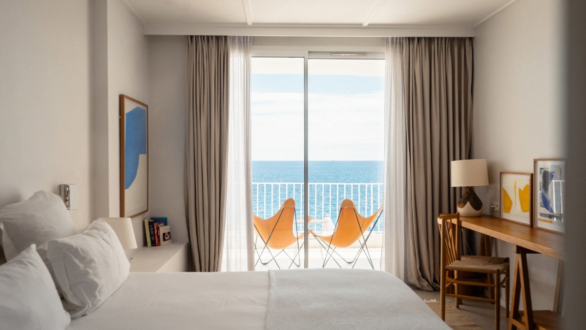 Bright hotel room with a bed, desk, and balcony featuring two orange chairs overlooking the ocean under clear skies.