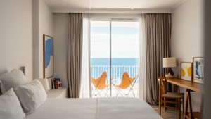 Bright hotel room with a bed, desk, and balcony featuring two orange chairs overlooking the ocean under clear skies.
