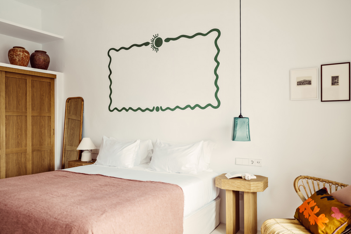 Chambre lumineuse avec couverture rose, art mural vert, fauteuil en osier et meubles en bois chaleureux.