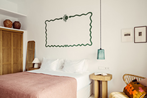 Chambre lumineuse avec couverture rose, art mural vert, fauteuil en osier et meubles en bois chaleureux.