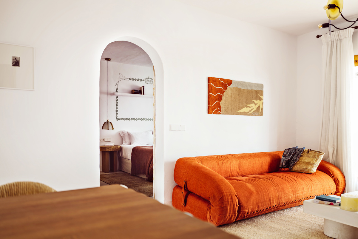 Salon lumineux avec un canapé orange, un coussin tissé, des œuvres murales, une table en bois et une vue sur la chambre.