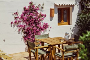 Terrasse extérieure avec table en bois, chaises vertes, bougainvillier rose sur mur blanc, et pichet en céramique.