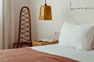 Coin chambre douillet avec lit blanc, chaise en rotin, table de chevet, lampe suspendue ambre, livre et tasse.