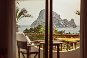 Vue depuis la chambre de deux transats sur terrasse surplombant l'océan et île rocheuse sous ciel.