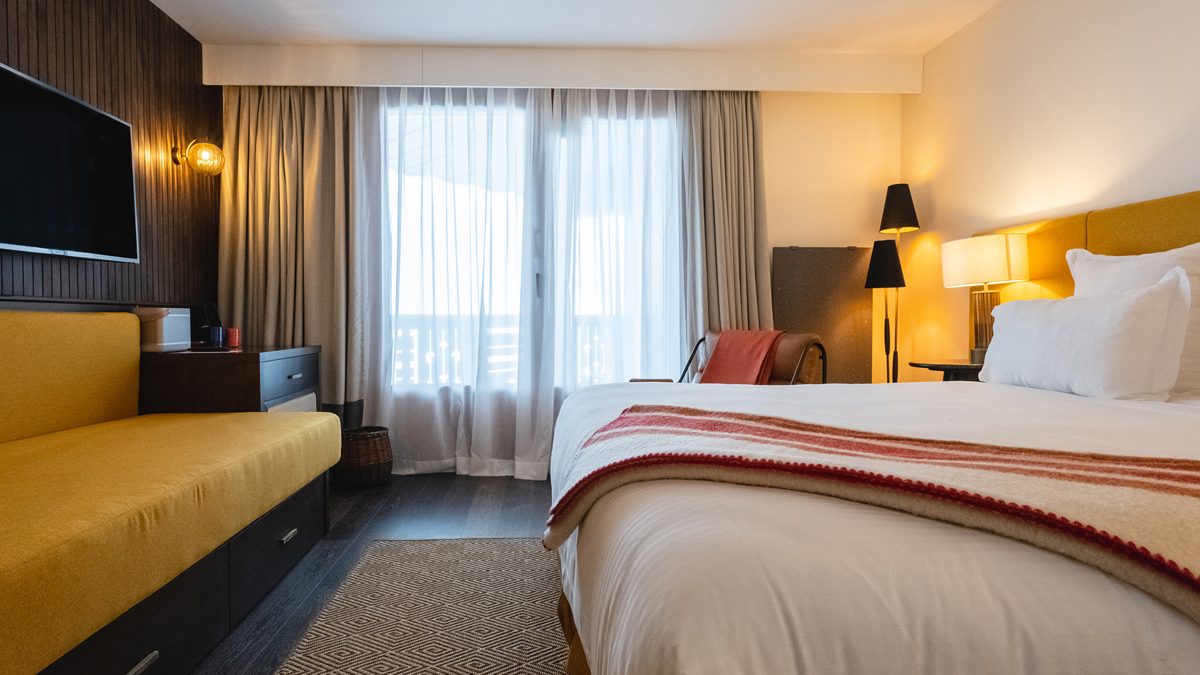 A bedroom features a TV mounted in front of the cozy bed and a stand lamp.