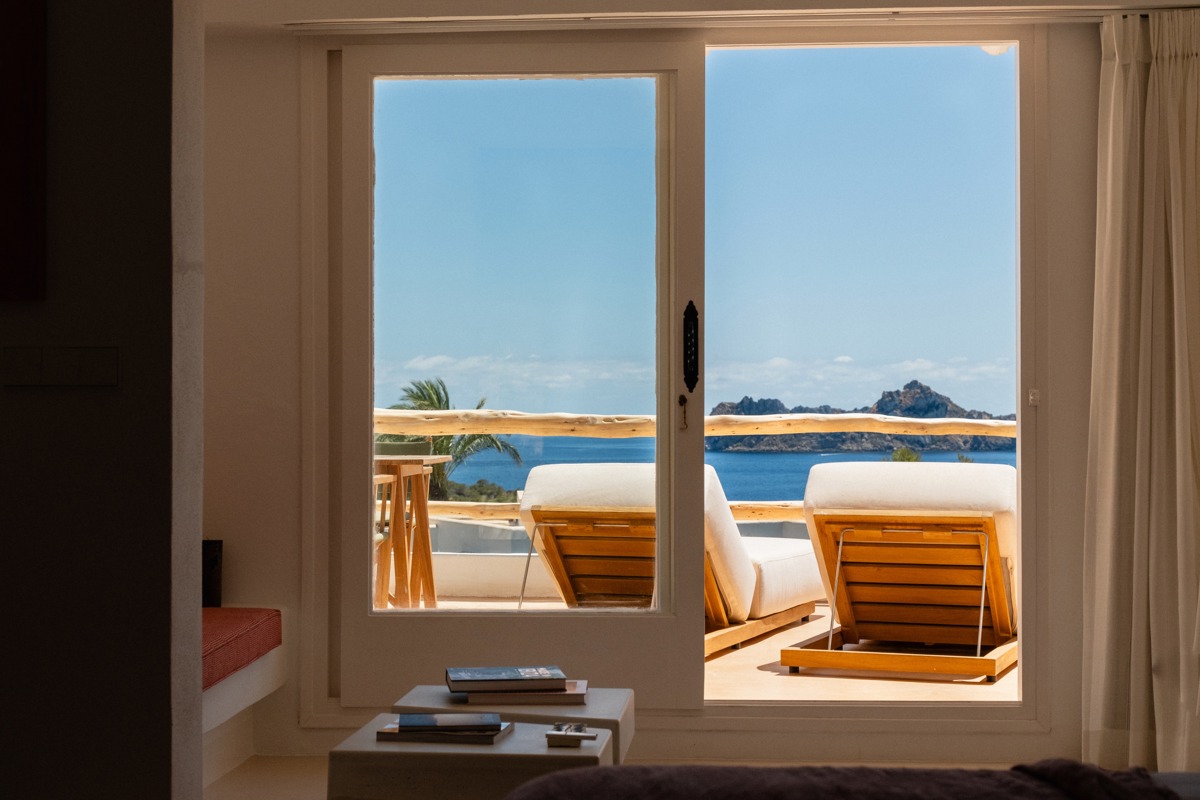 Vue à travers des portes vitrées sur des transats en terrasse surplombant la mer et îles rocheuses au loin.