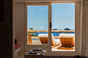 Vue à travers des portes vitrées sur des transats en terrasse surplombant la mer et îles rocheuses au loin.