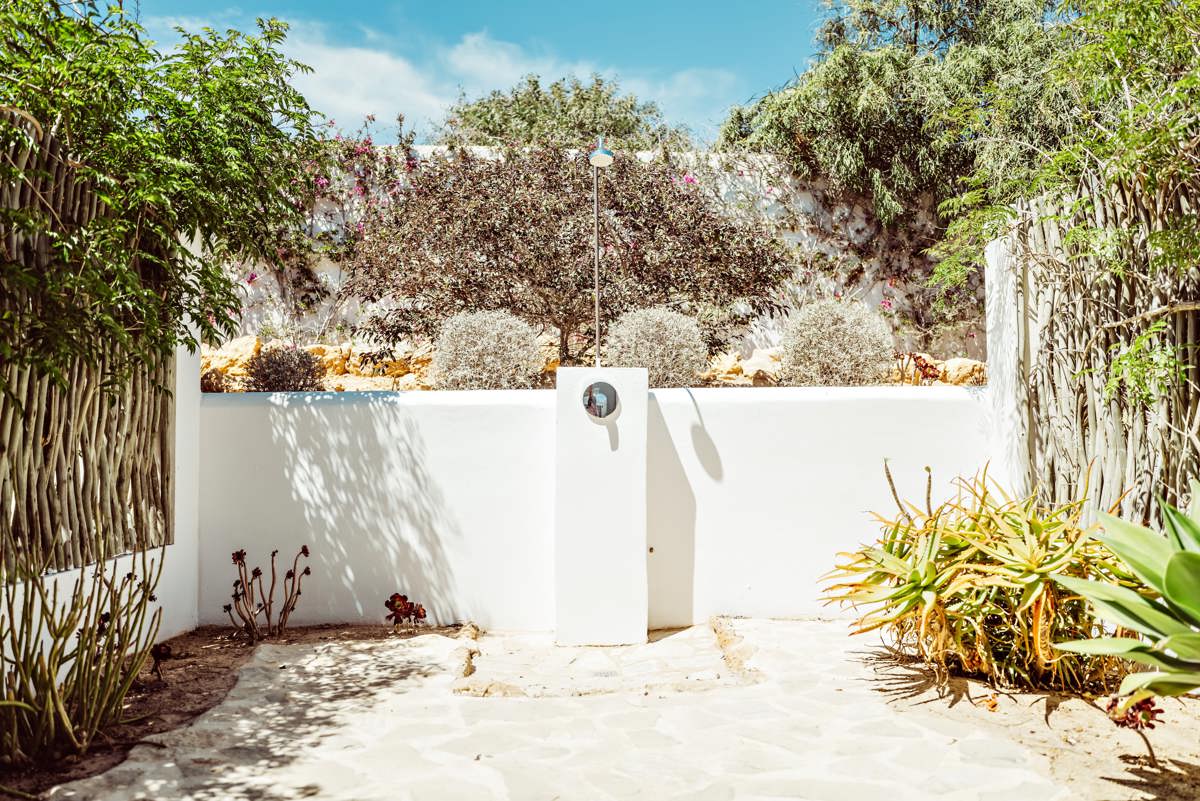 Douche extérieure en pierre blanche entourée de plantes luxuriantes, clôture rustique et ciel bleu éclatant.