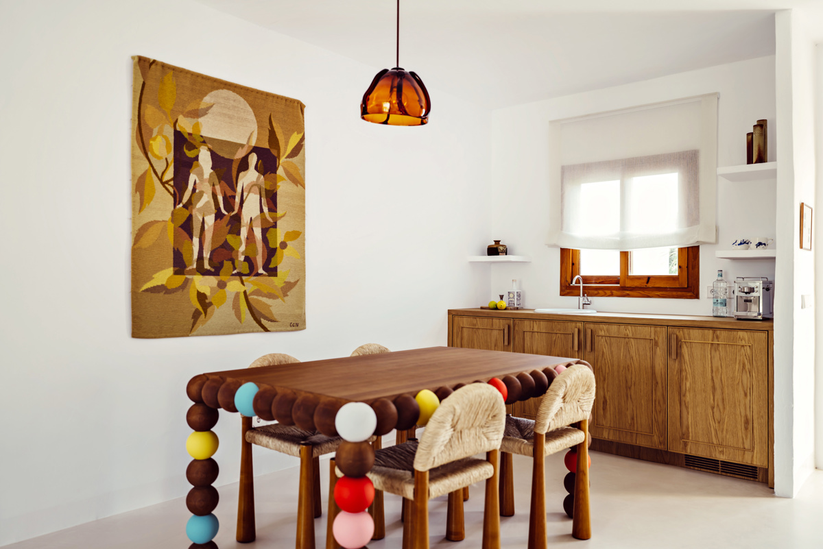 Cuisine lumineuse avec armoires en bois, table colorée, chaises tressées et tapisserie représentant des figures humaines.