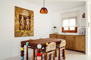 Cuisine lumineuse avec armoires en bois, table colorée, chaises tressées et tapisserie représentant des figures humaines.