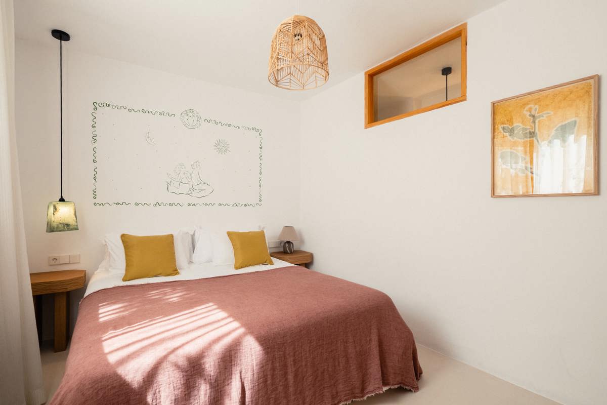 Chambre minimaliste et chaleureuse avec coussins moutarde, couvre-lit rose, dessin mural et suspension naturelle.