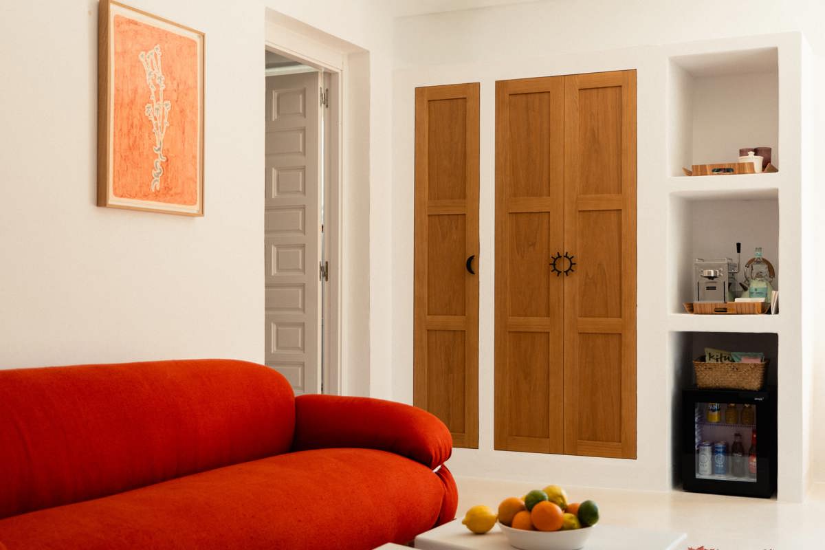 Espace de vie avec un canapé orange vif, une armoire en bois, des œuvres murales, un minibar intégré et une table avec des fruits.