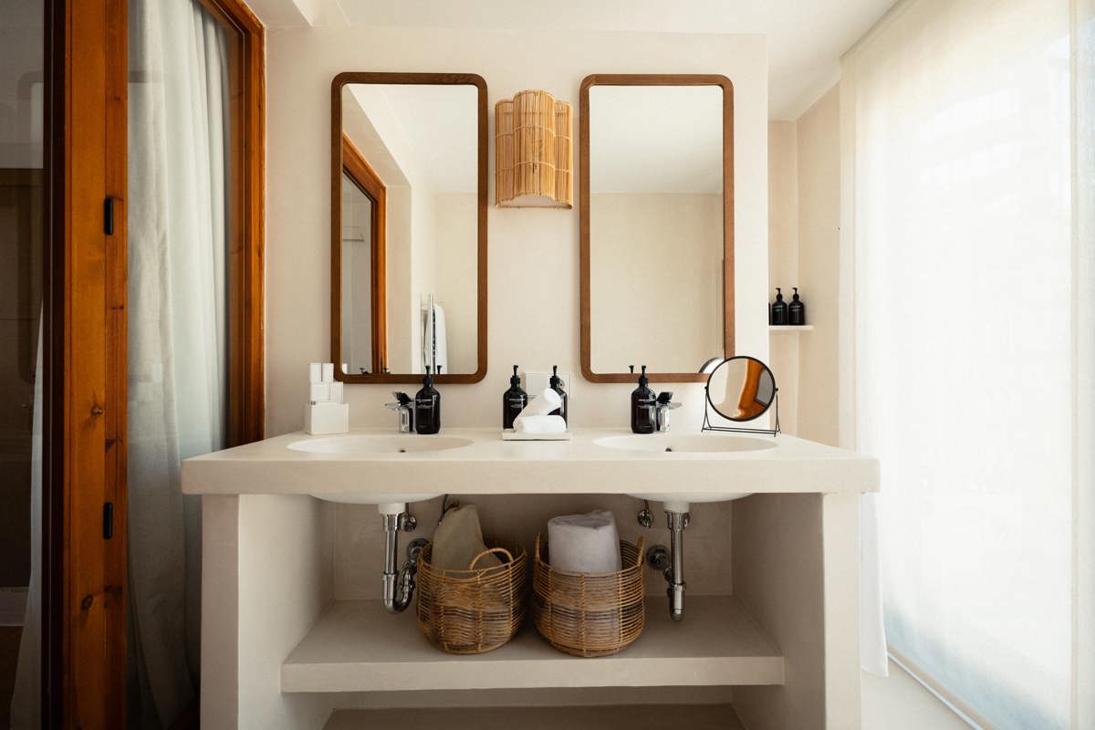Minimalist bathroom with double sinks, wooden mirrors, wicker baskets, and natural light through sheer white curtains.