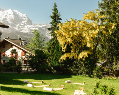 Outdoor yoga setup on a grassy garden lawn with mats and blocks, surrounded by trees and snow-capped mountains at Grand Hotel Belvedere in Wengen.