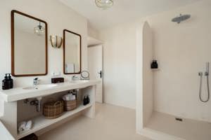 Bathroom with double sinks, large mirrors, open walk-in shower, wicker baskets, and neutral beige tones.