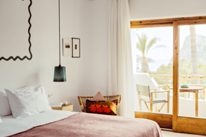 Chambre lumineuse avec un lit blanc, un plaid rose, une chaise en osier, une lampe suspendue et une vue depuis le balcon sur des palmiers.