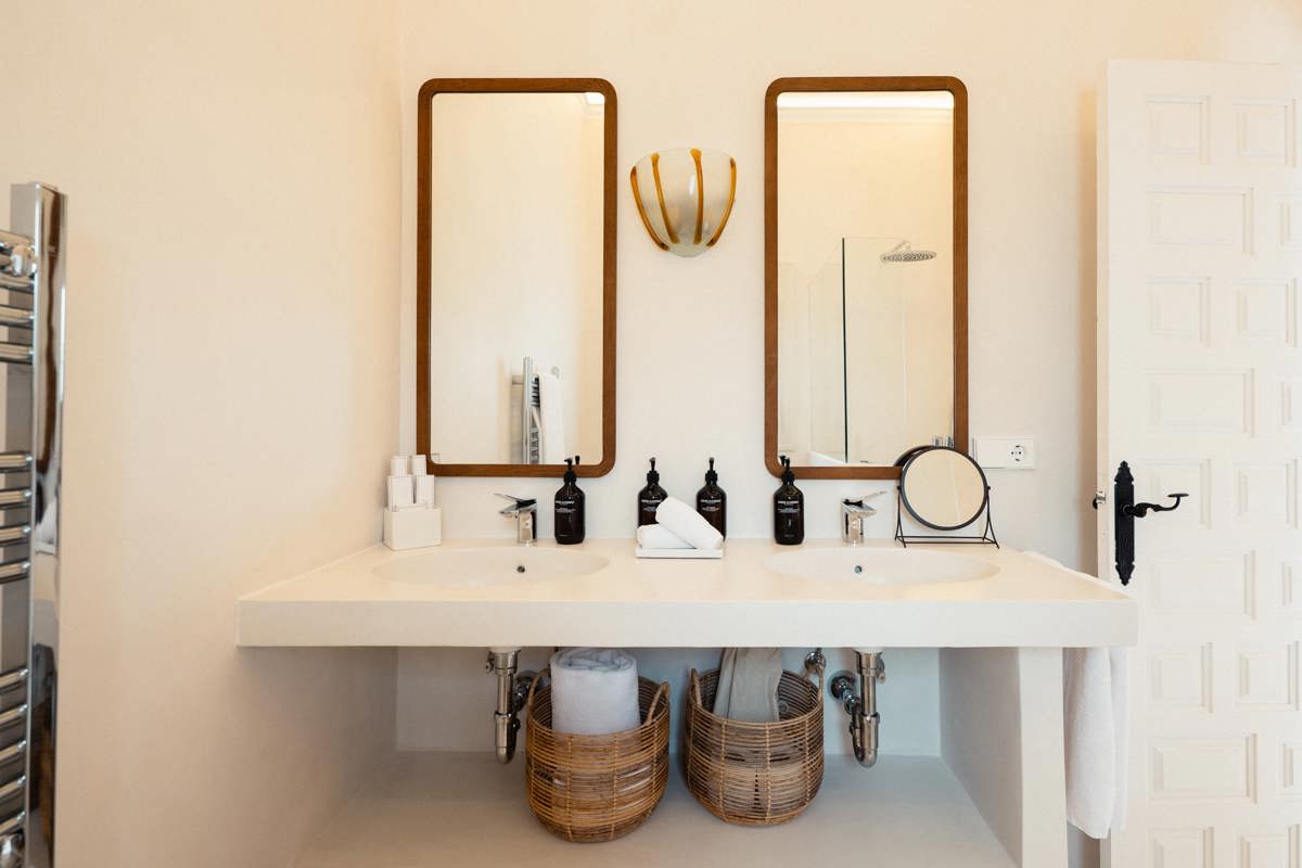 Salle de bain lumineuse avec double vasque, deux miroirs encadrés en bois, paniers en osier et porte blanche entrouverte.