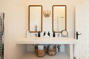 Salle de bain lumineuse avec double vasque, deux miroirs encadrés en bois, paniers en osier et porte blanche entrouverte.