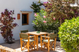 Terrasse ensoleillée avec table en bois, chaises vertes, fleurs roses et maison blanche méditerranéenne.