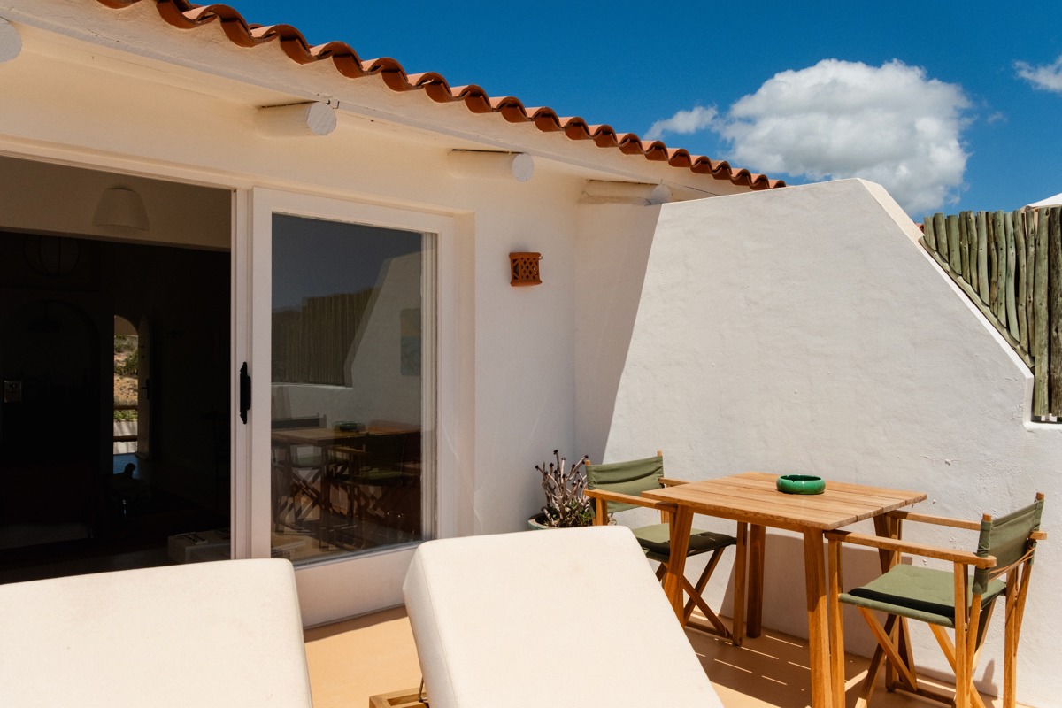 Terrasse ensoleillée avec transats blancs, table en bois, chaises vertes et mur blanc sous un ciel bleu clair.