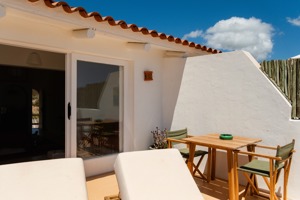 Terrasse ensoleillée avec transats blancs, table en bois, chaises vertes et mur blanc sous un ciel bleu clair.