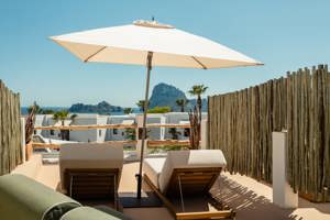 Terrasse ensoleillée avec transats sous un parasol blanc, vue sur mer bleue et îles rocheuses.