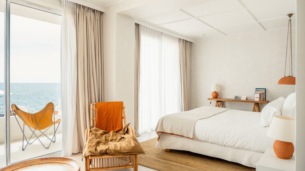 A tidy bedroom features a cozy bed, a nearby chair, and a balcony chair offering a wonderful ocean view.