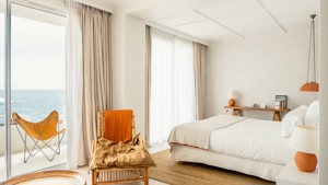 A tidy bedroom features a cozy bed, a nearby chair, and a balcony chair offering a wonderful ocean view.