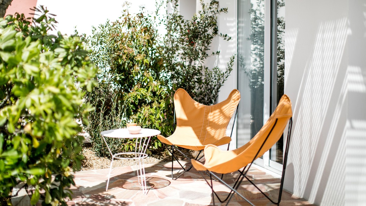 Sunny patio with two orange chairs, a white table, and lush greenery.