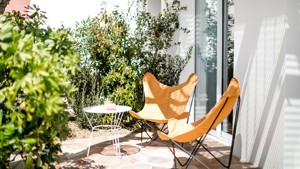 Sunny patio with two orange chairs, a white table, and lush greenery.
